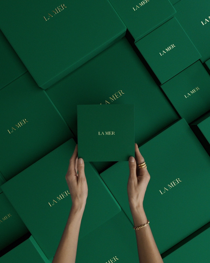 Hands with white gloves placing lid on green giftbox, then tying a gold la mer ribbon into a bow, cutting ends of ribbon.